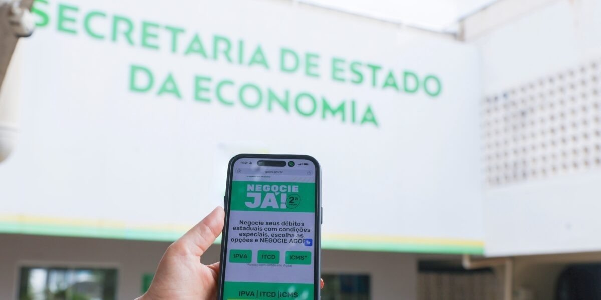 Celular com a página do Negocie Já II aberta diante da fachada da Secretaria de Estado da Economia de Goiás