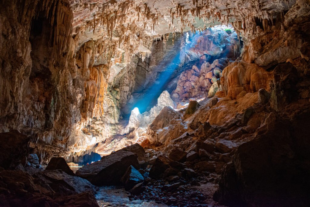 Interior da caverna Terra Ronca, em São Domingos (GO), com feixe de luz azul entrando pelo teto, estalactites e rio no fundo
