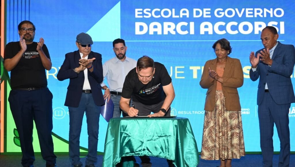 Prefeito Sandro Mabel assina documento no lançamento da Escola de Governo Darci Accorsi, em Goiânia, aplaudido por autoridades, com painel azul ao fundo