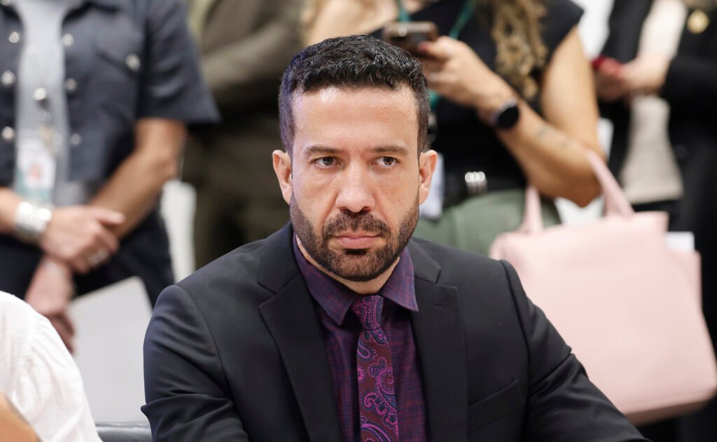 Deputado federal André Janones sentado em reunião na Câmara dos Deputados, de terno escuro e gravata roxa
